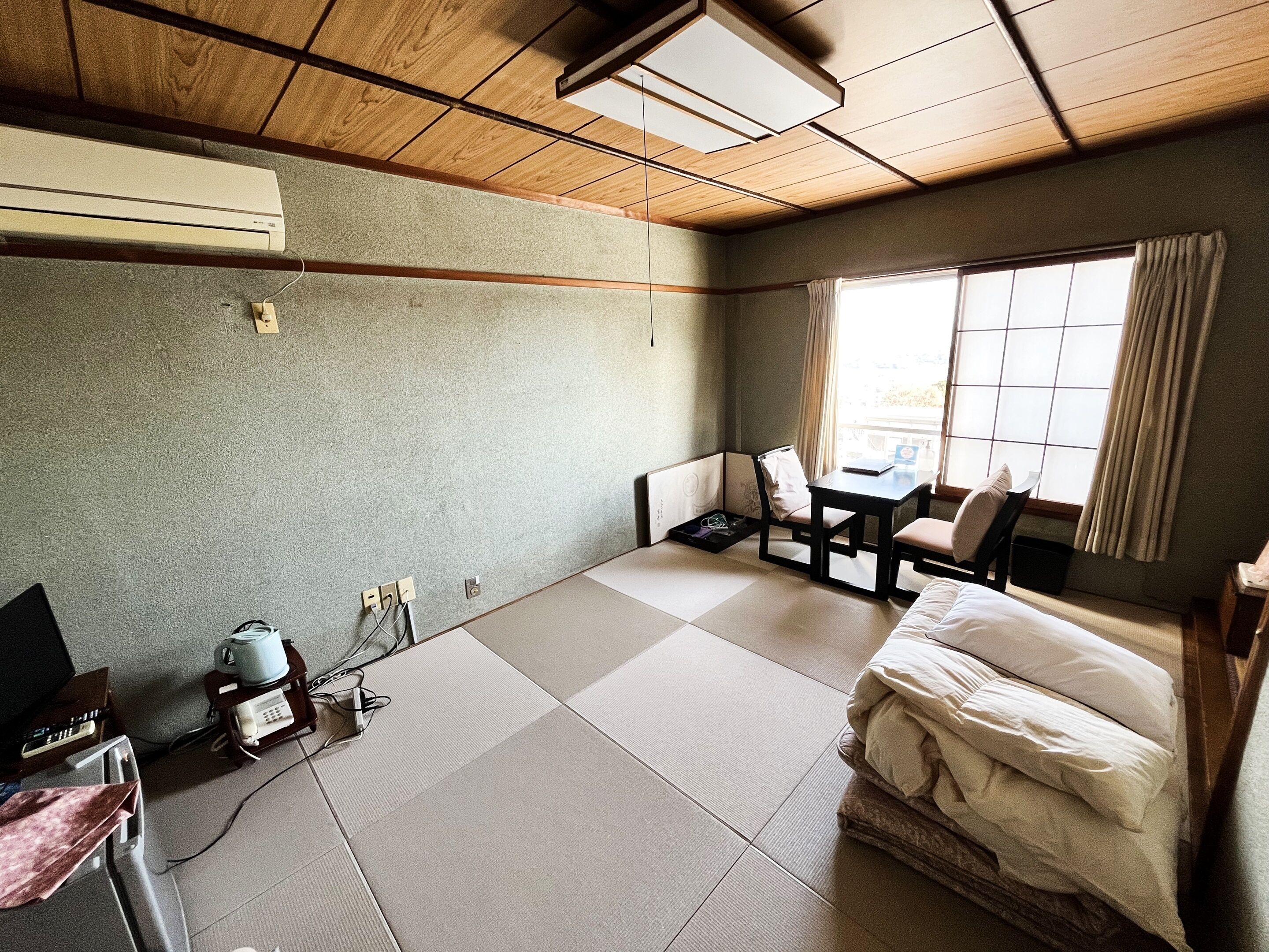 A room with tatami flooring, a bed with white bedding, a small table, a chair, and a window with curtains.