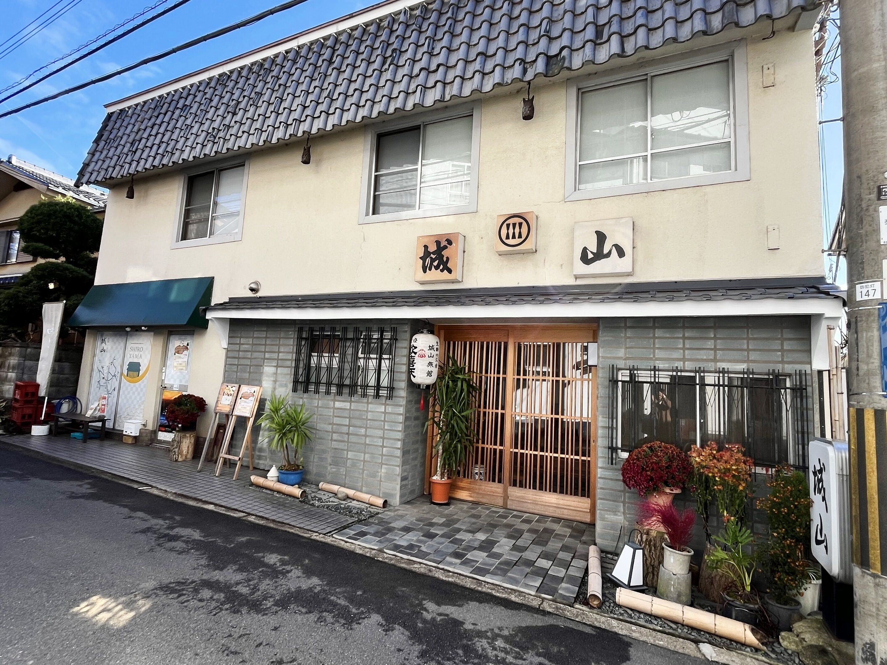 Um edifício japonês tradicional com telhado de telhas, uma placa com caracteres japoneses e uma pequena entrada com portão de madeira.