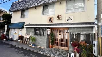 Um edifício japonês tradicional com telhado de telhas, uma placa com caracteres japoneses e uma pequena entrada com portão de madeira.