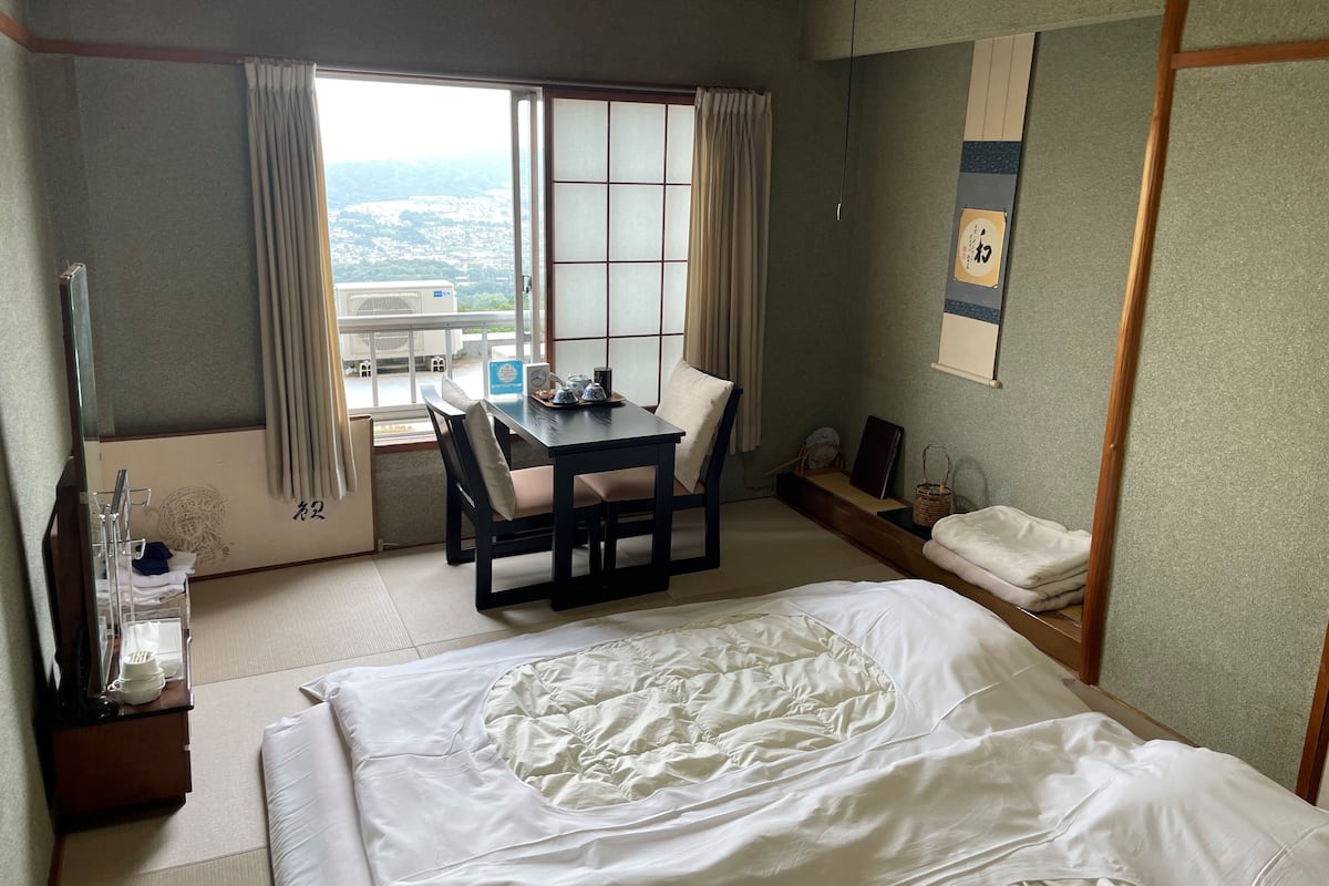 A hotel room with a bed, a desk with a chair, a window with curtains, and a view of the city.