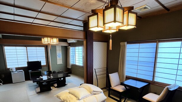 A traditional Japanese-style room with tatami flooring, a futon bed, a low table with chairs, a television, and a large window with shoji screens. - Shiroyama inn (Ikoma)