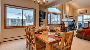 A dining area with a wooden table set for four, large windows, a fireplace, and a cozy seating area with a leather couch.