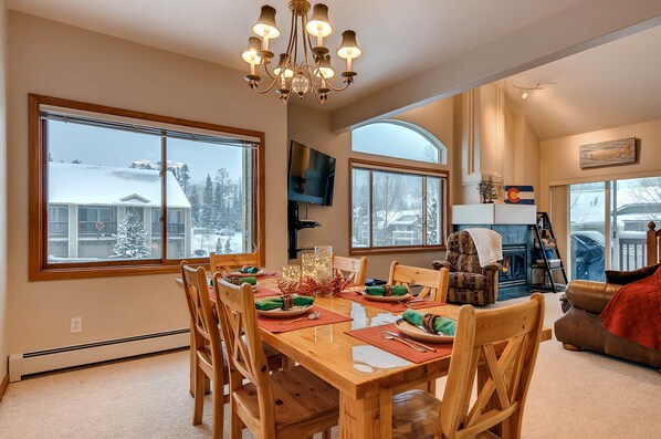 A dining area with a wooden table set for four, large windows, a fireplace, and a cozy seating area with a leather couch.