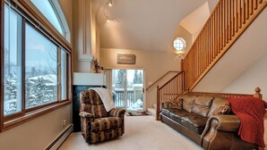 A cozy living room with a fireplace, a leather sofa, a plaid armchair, and a wooden staircase.