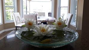 A dining area with a glass table centerpiece featuring water lilies, white chairs, and a table with a floral tablecloth. - water front luxury lake house (Janetville)