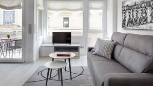 Uma sala de estar moderna com um sofá cinzento, uma televisão de ecrã plano, uma mesinha com um livro e vista para a cidade através de grandes janelas.