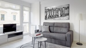 Uma sala de estar moderna com um sofá cinzento, uma televisão de tela plana sobre um móvel branco, uma mesa redonda pequena com um livro e uma grande pintura em preto e branco retratando uma paisagem urbana na parede.