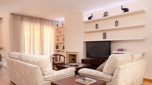 A living room with two patterned armchairs, a wooden coffee table, a flat-screen TV, and shelves with decorative items.