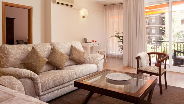 A modern living room with a sofa, a glass coffee table, and a chair. There is a small table with a laptop and a plant.