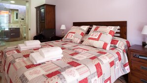 A neatly made bed with a red and white quilt, a wooden nightstand with a lamp, and a bathroom visible in the background.