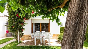 A patio with two white plastic chairs and a table, surrounded by lush greenery and a tree. - INNOUTHOME HOUSE WITH GARDEN AND SWIMING-POOL, BBQ AC AND WIFI,_JUAN DE ALMENARA (L'AMETLLA DE MAR)
