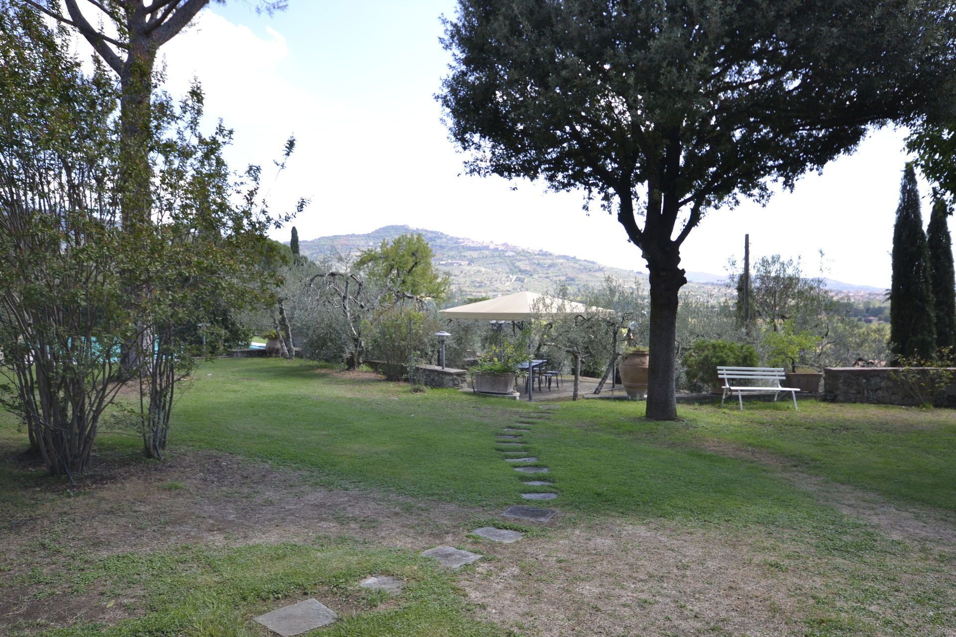 Un jardin avec un sentier en pierre menant à un espace aménagé pour s’asseoir, entouré d’arbres et offrant une vue sur des collines au loin.