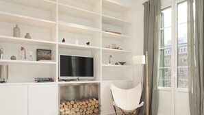 A modern living room with white shelving, a flat-screen TV, a fireplace, and a view of buildings through a large window.
