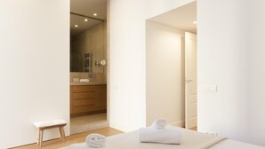 A modern bedroom with a large bed, two rolled towels, a small stool, and a bathroom visible through an open door.