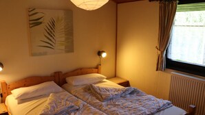 Una habitación con cama doble, dos mesitas de noche, una lámpara de pared, una ventana con cortinas y una planta en la pared.