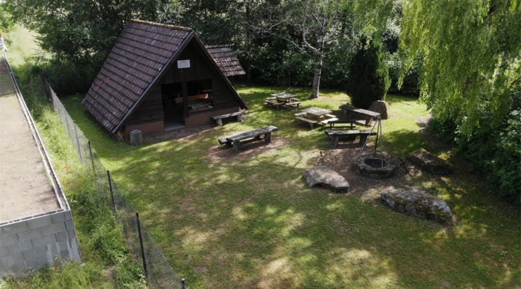 A rustic cabin with a sloped roof, surrounded by a fenced yard with picnic tables and a fire pit.