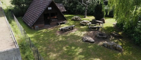A rustic cabin with a sloped roof, surrounded by a fenced yard with picnic tables and a fire pit.
