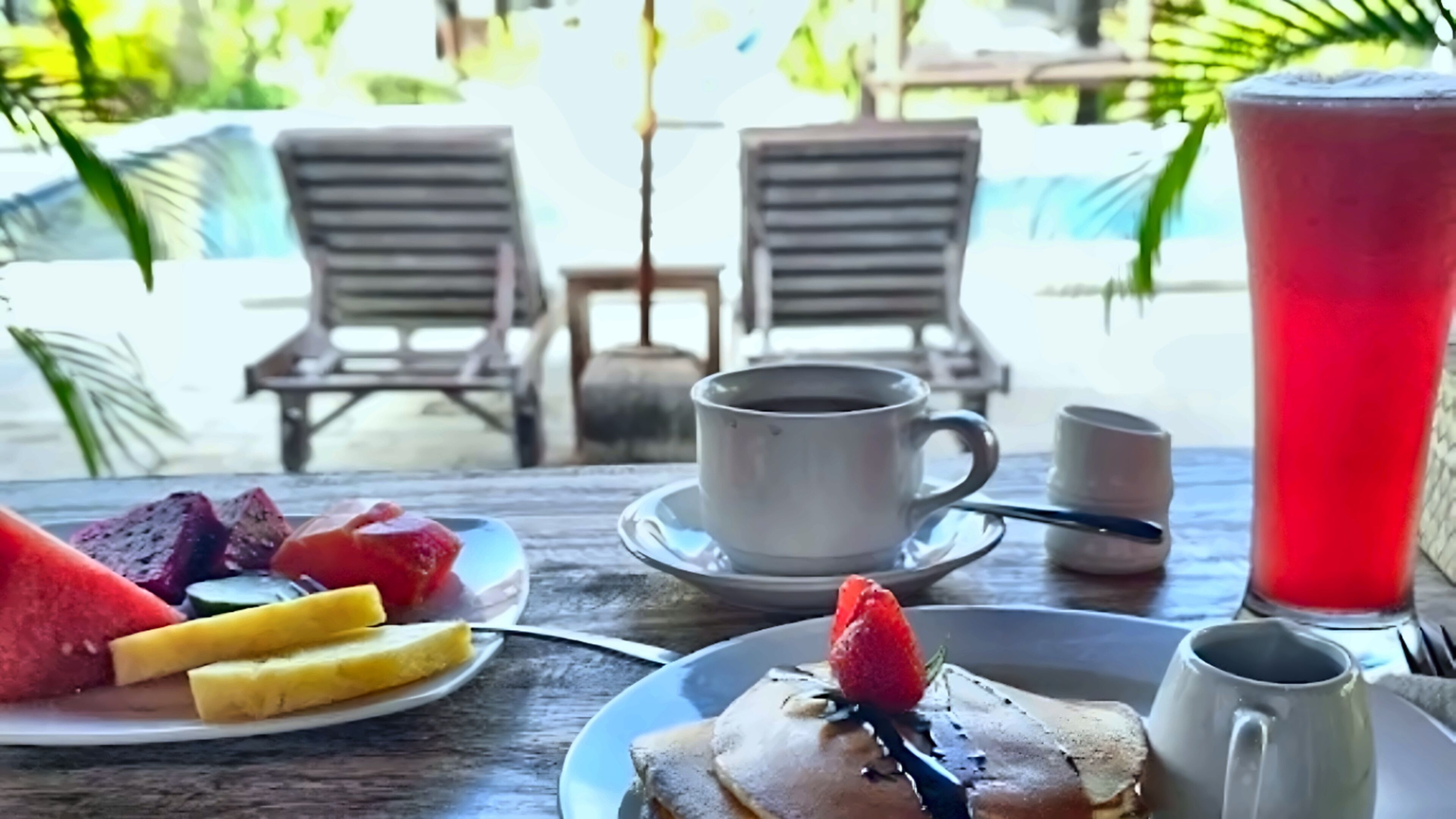 Un desayuno con un plato de fruta, panqueques y una bebida roja, con vista a una piscina y sillones de descanso al fondo.