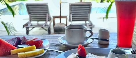 Un desayuno con un plato de fruta, panqueques y una bebida roja, con vista a una piscina y sillones de descanso al fondo.