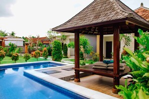 Un área de piscina con un quiosco de madera, rodeado de follaje y un cielo despejado.