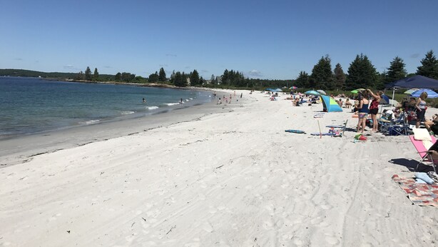 Uma praia com céu claro, areal arenoso e pessoas relaxando sob guarda-sóis.