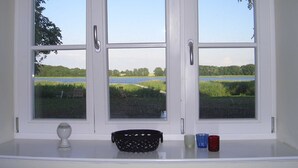 Een raam met drie ruiten, een witte plank eronder, en uitzicht op een meer en groen.