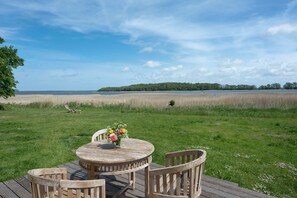 A wooden deck with a round table and chairs, overlooking a grassy area with a body of water and a distant tree line.