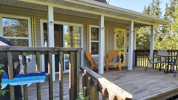 A wooden deck with a covered porch, outdoor seating, and a sign reading "Little Eden Summer Beach Home." - Little Eden Summer Beach Home  (Canoe Cove)
