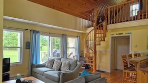 A spacious living room with a grey sofa, wooden staircase, and a dining area with wooden chairs. - Little Eden Summer Beach Home  (Canoe Cove)