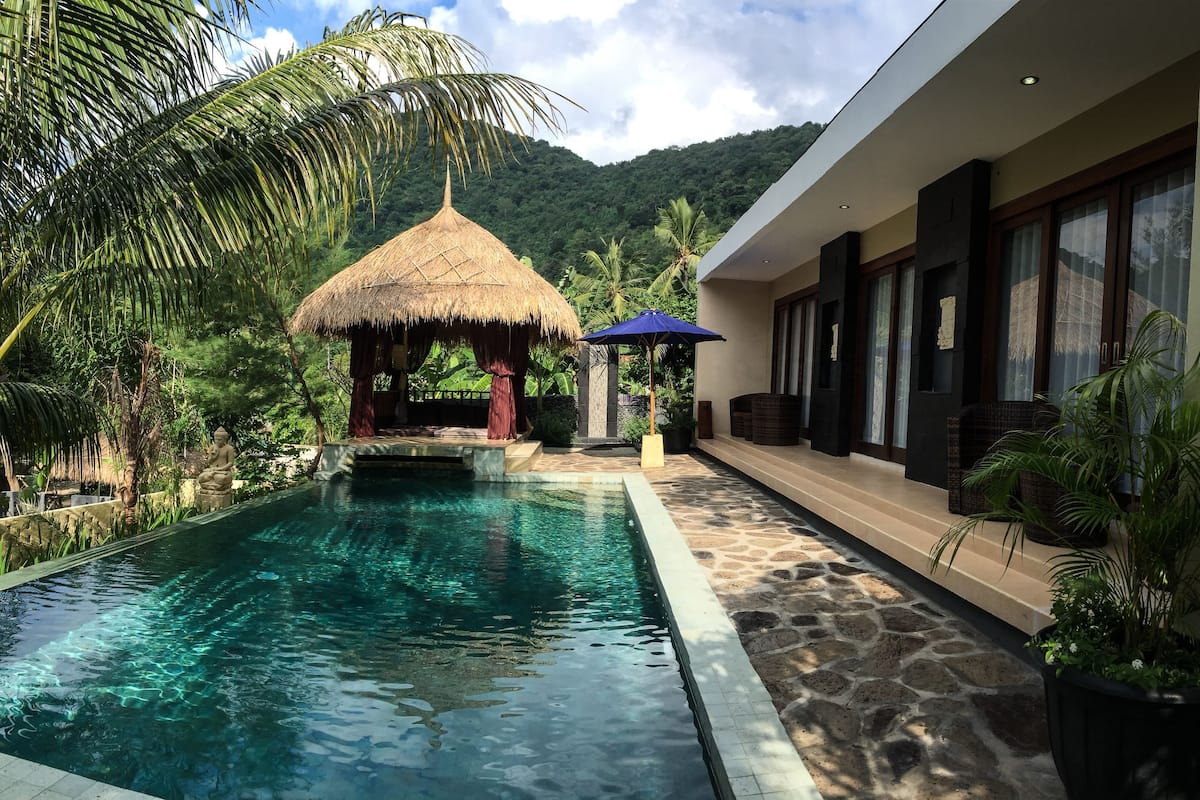 A pool area with a thatched-roof structure, a stone pathway, and a modern building with large windows.
