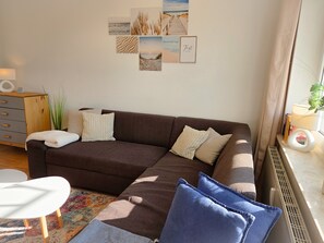 A living room with a brown sectional sofa, a white round table, and a colorful rug. There are wall art pieces above the sofa and a plant on the windowsill.