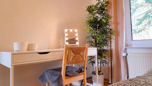 Quarto bem arrumado com cama, mesa de cabeceira, escrivaninha com espelho e planta em vaso.