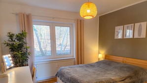 Quarto com cama, mesa de cabeceira com abajur, janela com cortinas, radiador e planta em vaso.