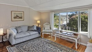 Ein gemütlicher Wohnraum mit einem gestreiften Sofa, einem gemusterten Teppich, einem weißen Sessel und einem kleinen Tisch mit Büchern. Ein gerahmtes Bild hängt an der Wand, und ein Fenster bietet Blick auf grüne Vegetation draußen.