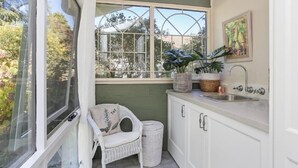 Eine kleine, abgeschlossene Küche mit Fenster, einem Korbstuhl, einem Spülbecken und einem Mülleimer.