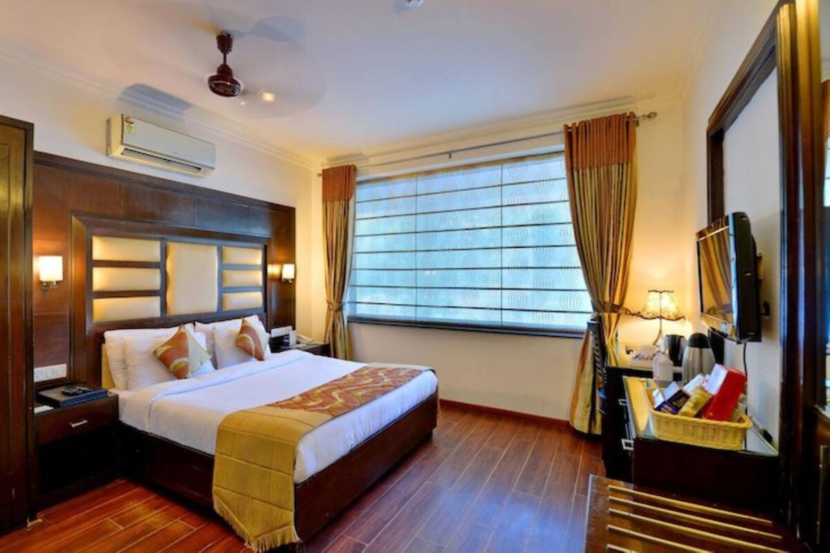 A hotel room with a large bed, a flat-screen TV, a desk with a lamp, and a window with curtains.
