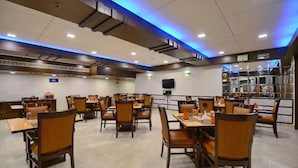 Een ruim restaurant met meerdere eettafels en stoelen, een flatscreen televisie aan de muur en een modern plafondontwerp met blauwe lichtaccenten.