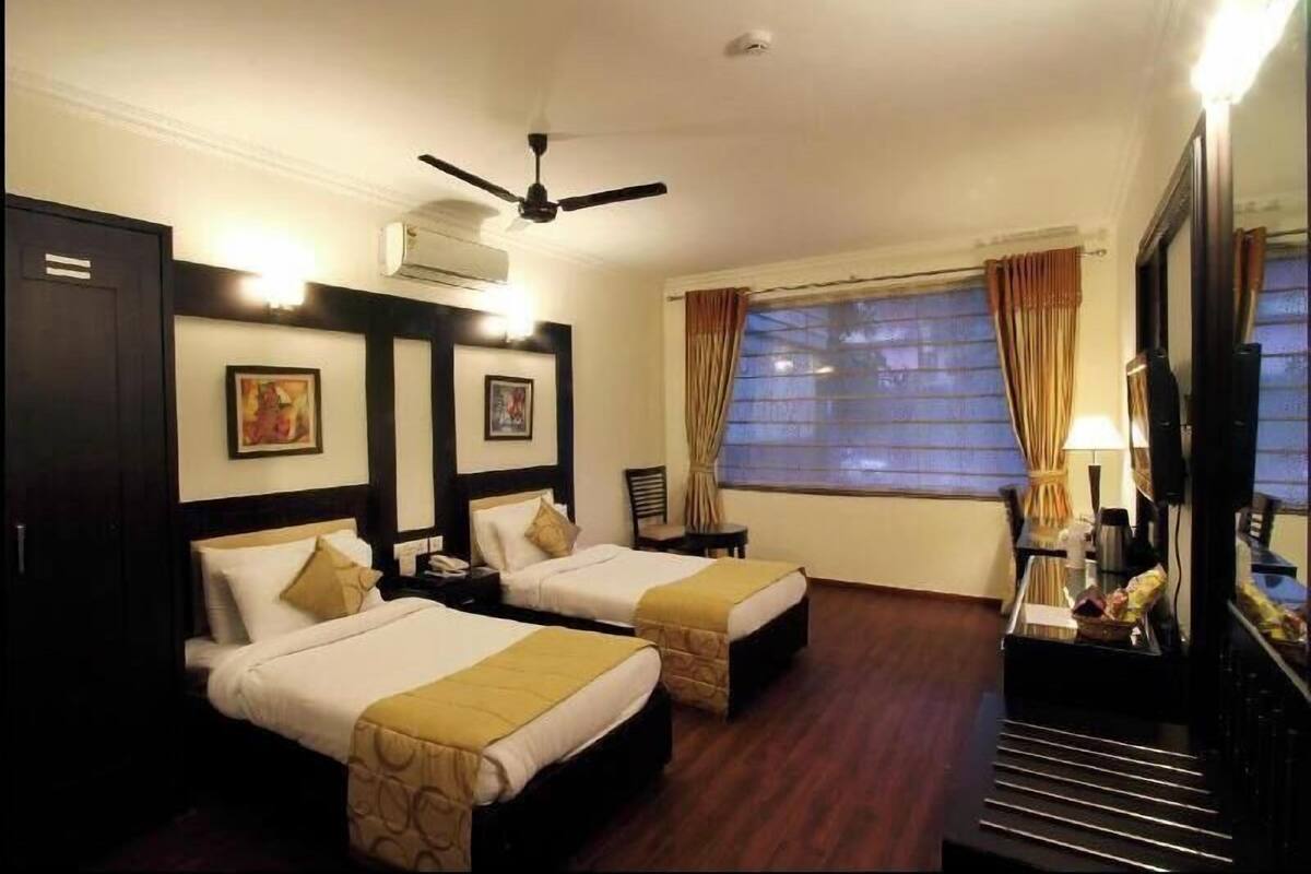 A hotel room with two beds, a ceiling fan, a large window with curtains, a TV, and a mirror.