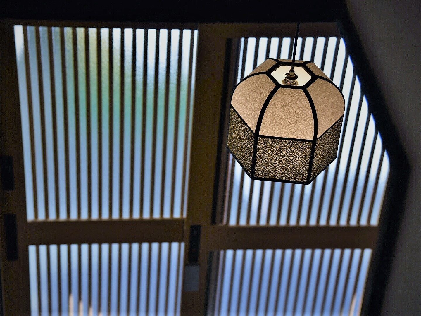 A hanging lantern with a geometric pattern, suspended in front of a window with vertical blinds.