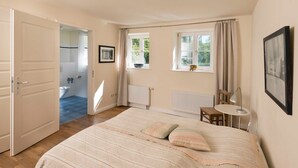 Une chambre à coucher avec un lit, un bureau, une chaise, une fenêtre avec des rideaux et une salle de bain visible à travers une porte ouverte.