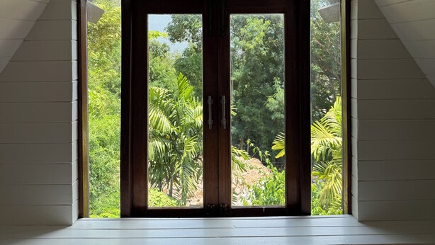 A room with a sloped ceiling, a large window with a view of greenery, and a wooden door. - Private Thai style villa with private  pool (Koh Samui)