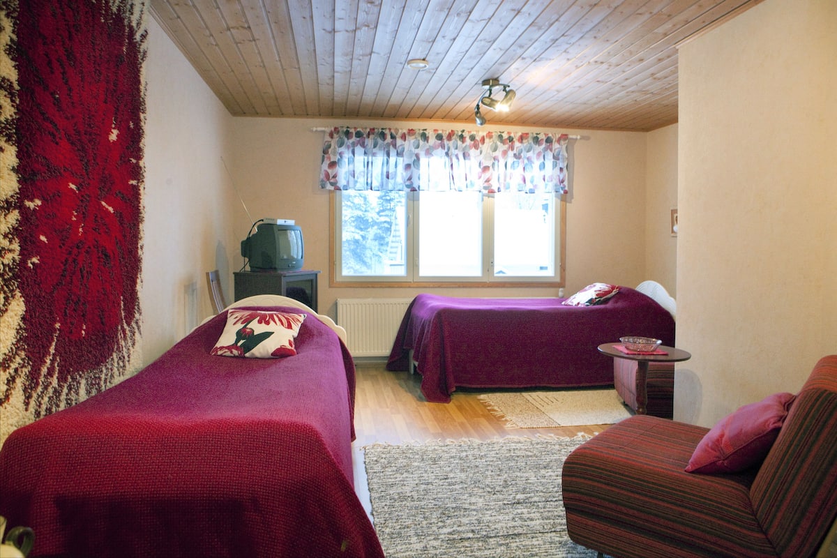 A room with two beds, a small table, a sofa, a television, and a window with curtains.