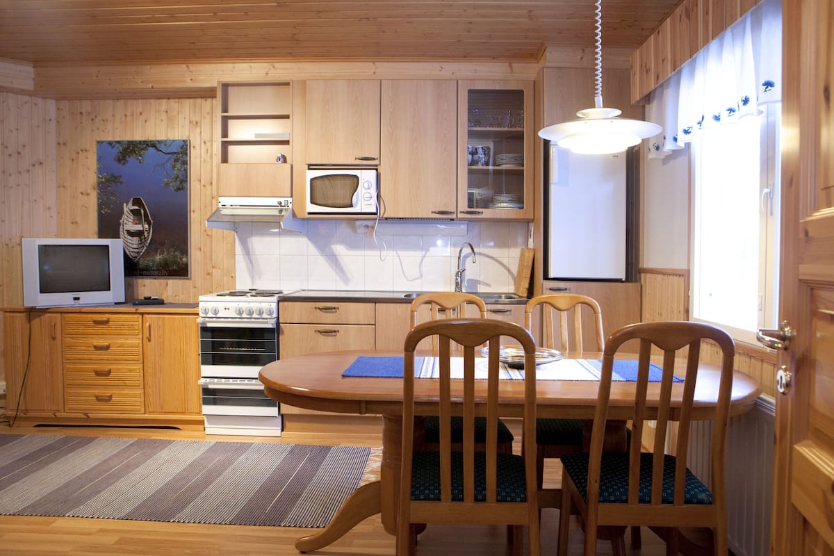 A wooden cabin-style kitchen with a dining table, chairs, a microwave, stove, and a television.