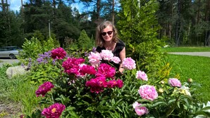 Une femme se tient au milieu d’un jardin où s’épanouissent de magnifiques fleurs, avec une voiture et des arbres en arrière-plan.