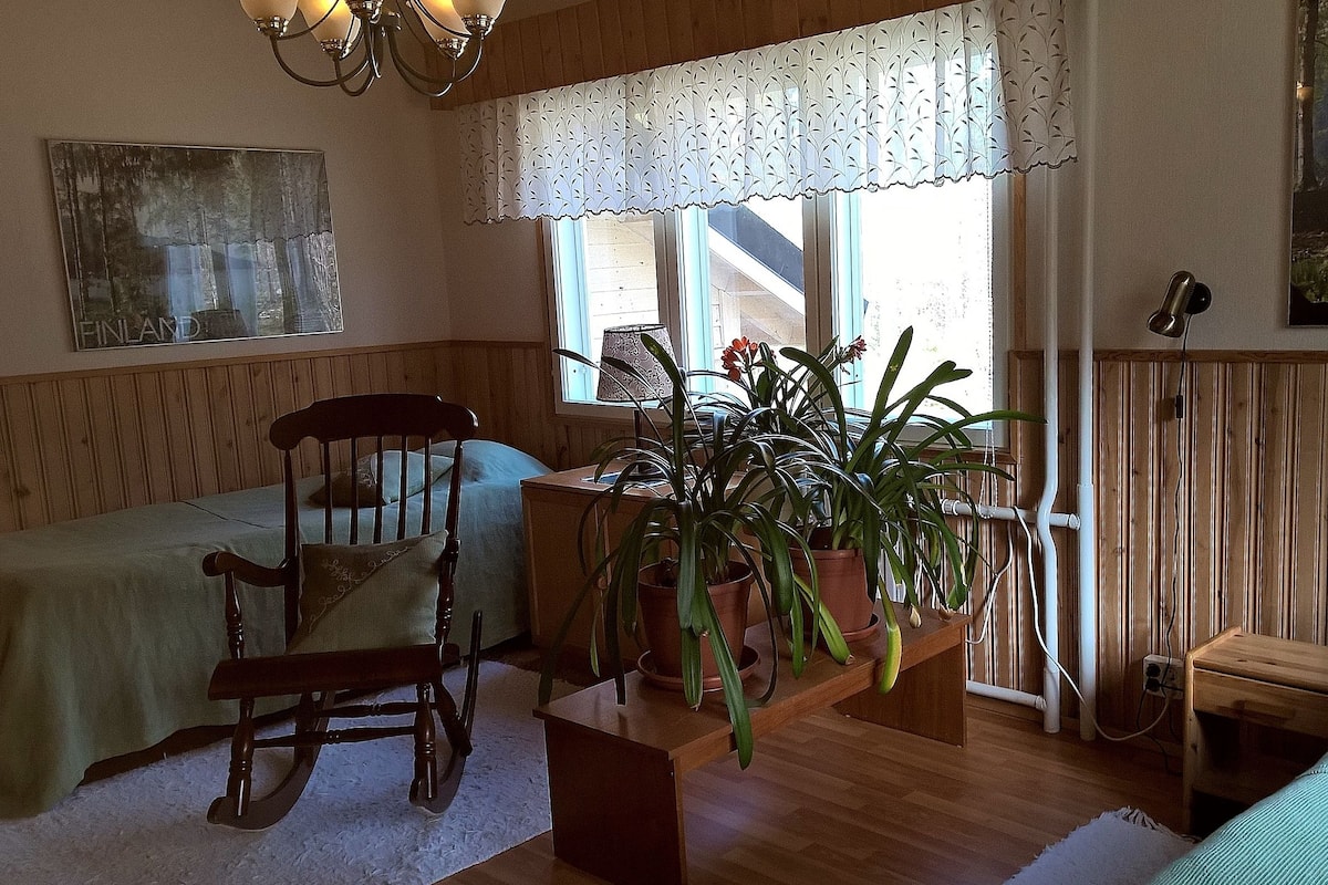 A cozy room with a rocking chair, a small table with a potted plant, a bed with a green bedspread, and a window with a lace curtain.
