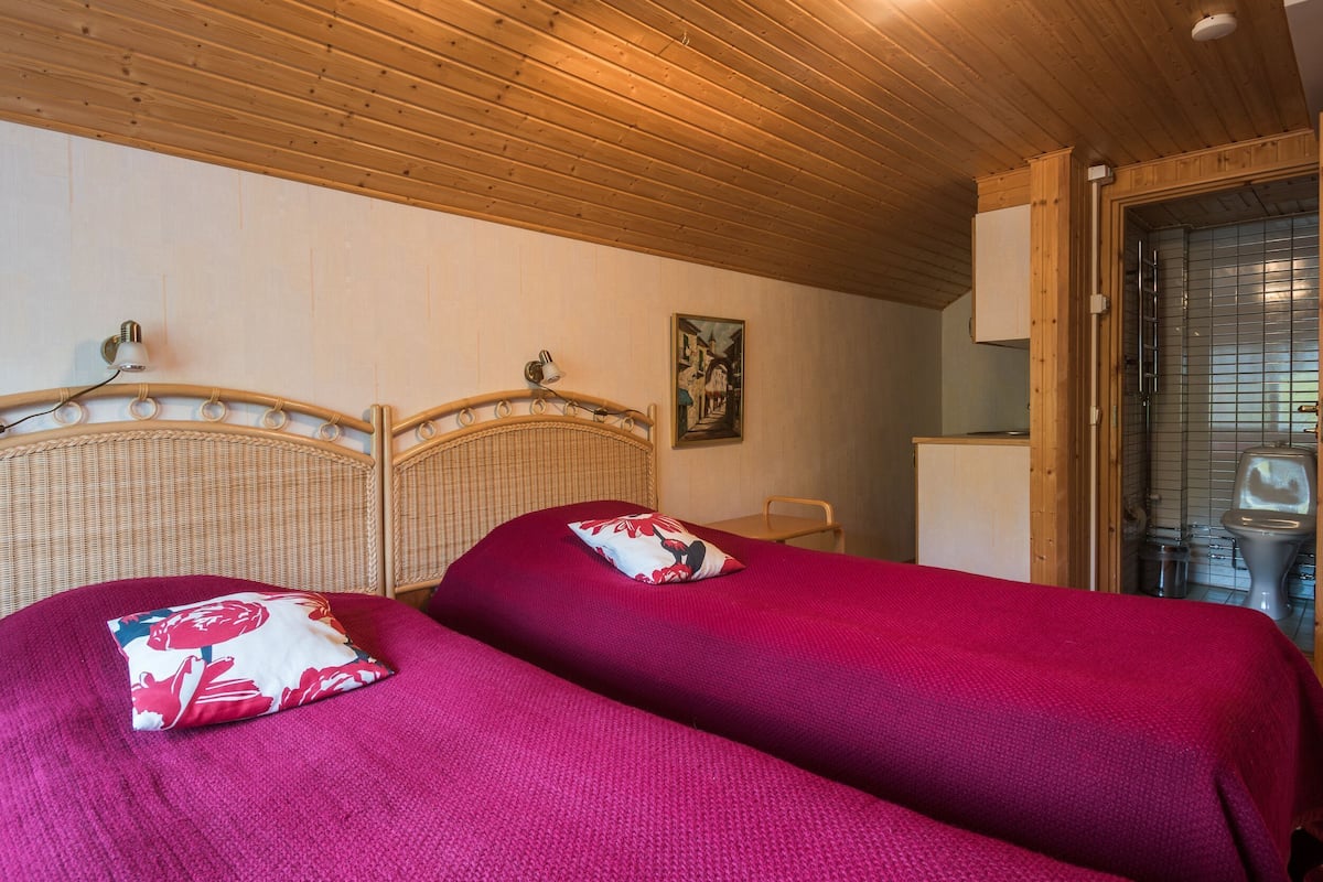 A room with two beds, wooden ceiling, a painting on the wall, and a bathroom visible through a glass door.