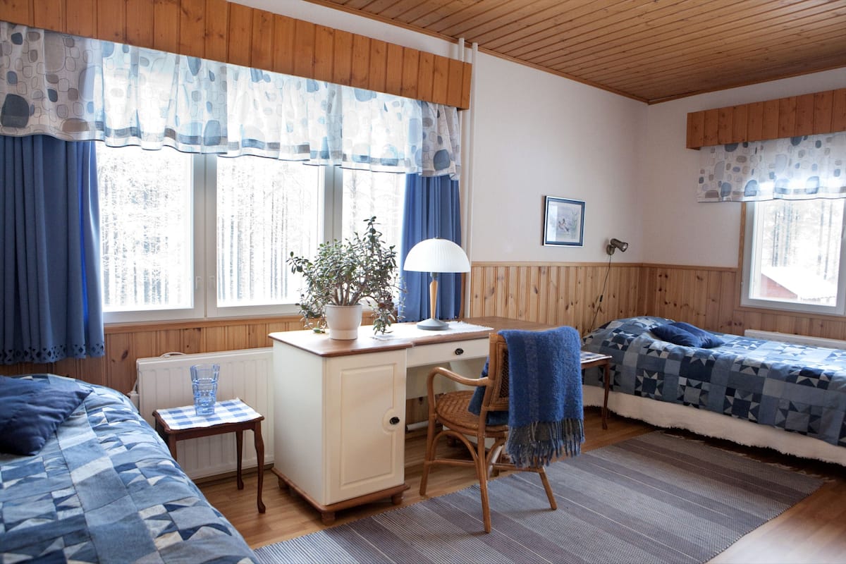 A room with a wooden ceiling, a desk with a lamp, a chair, a bed with a blue quilt, and a window with curtains.