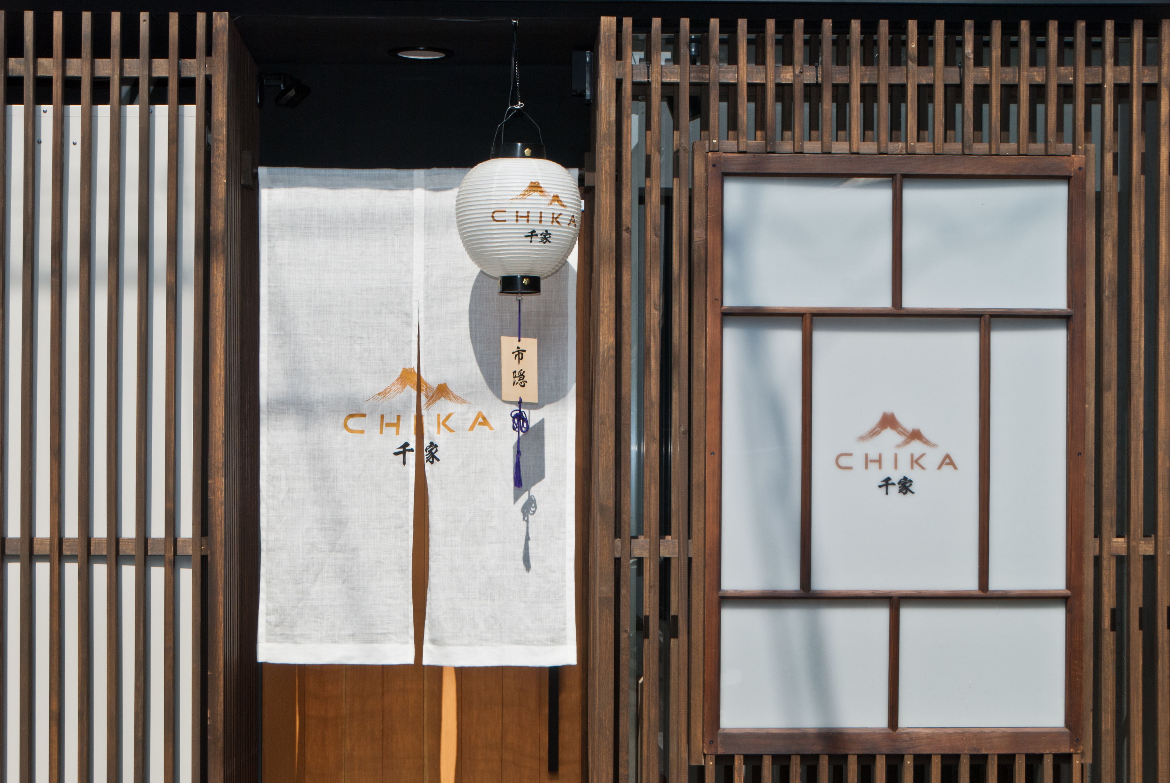 Una entrada japonesa tradicional con una puerta de celosía de madera, un farol colgante y una puerta corredera de papel con el logotipo de Chika.