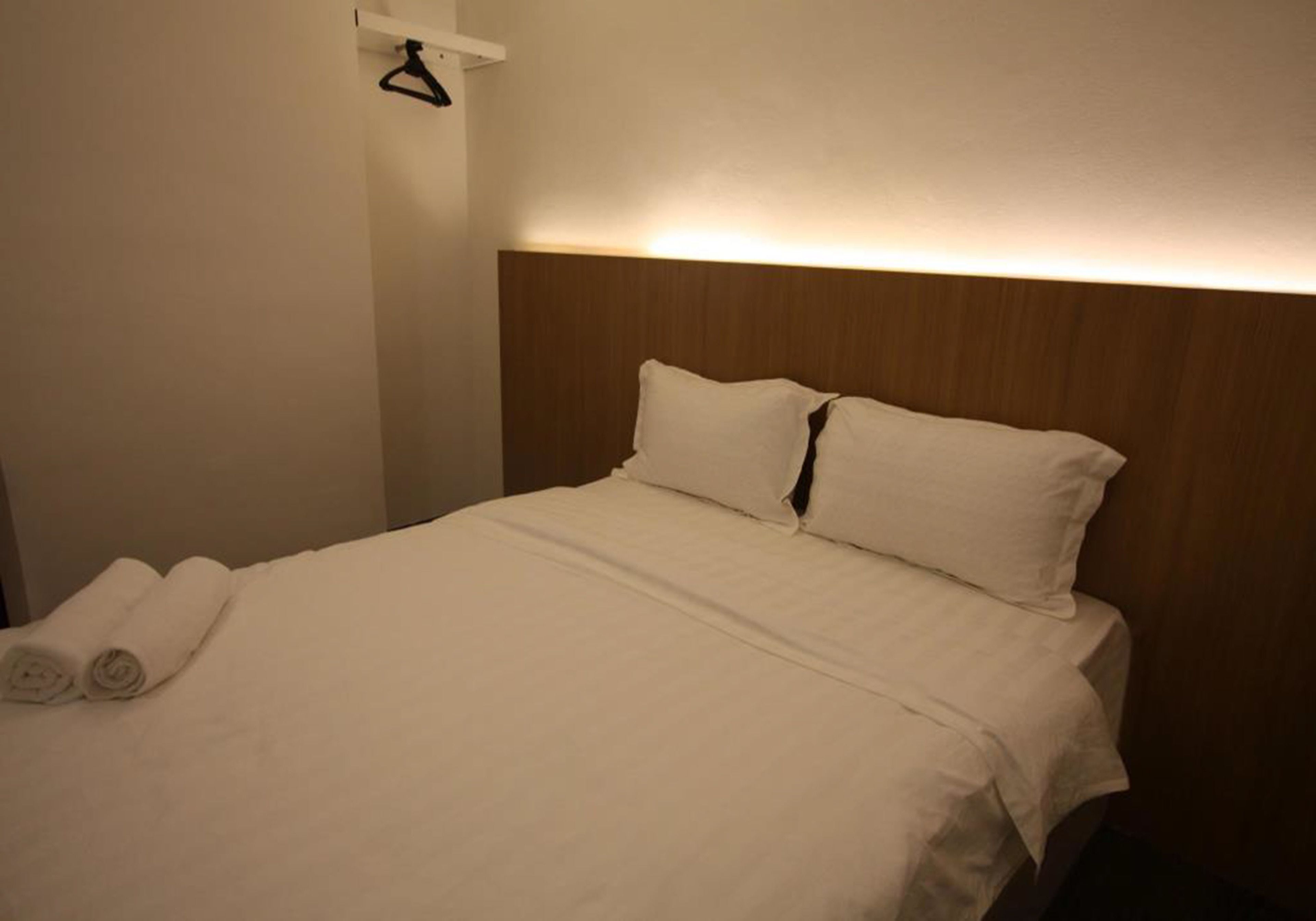 Una cama bien hecha con sábanas y almohadas blancas, un cabecero de madera y un perchero para toallas.
