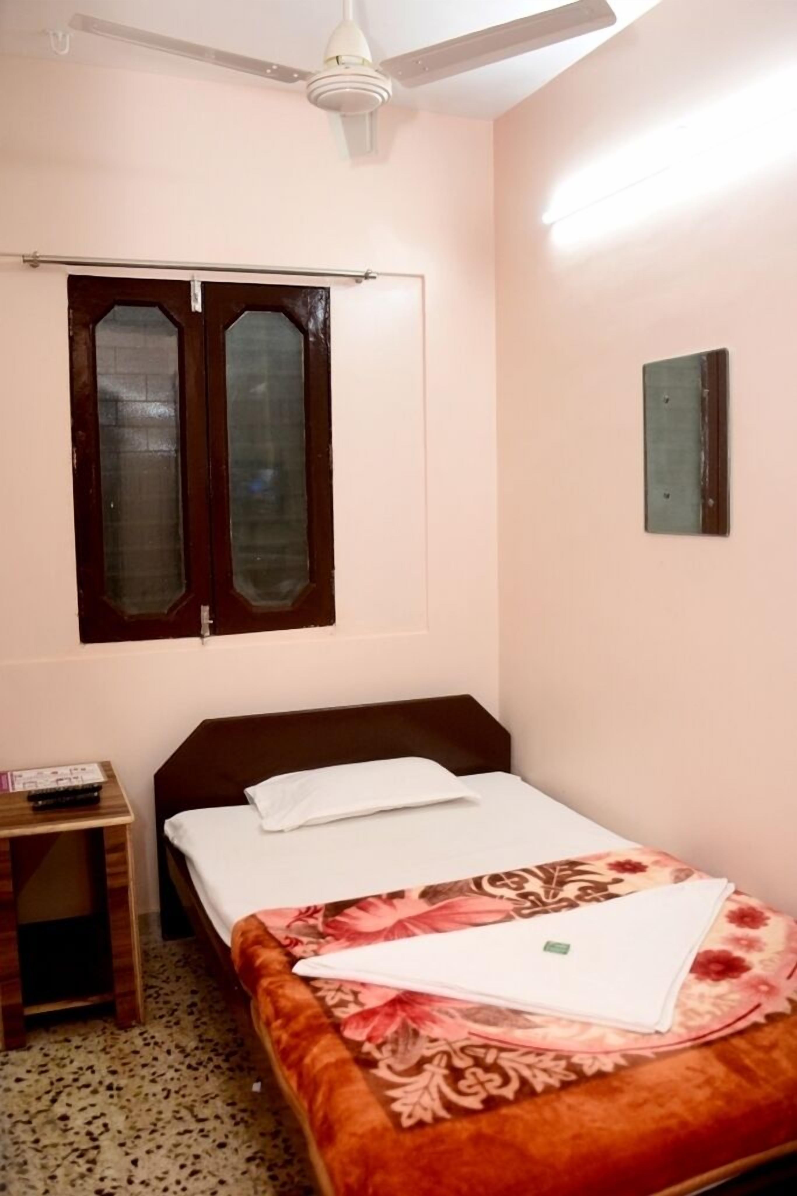 A bedroom with a bed, a nightstand, a window with curtains, a ceiling fan, and a wall-mounted light fixture.
